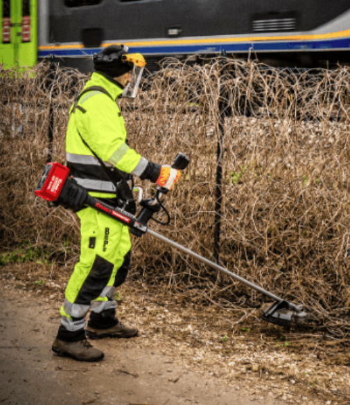 A worker in high-visibility safety gear uses a brush cutter to clear overgrown vines along a fence line as a train passes in the background