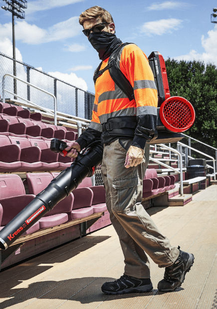 A worker in high-visibility safety gear operates a Kress Commercial backpack leaf blower while walking through rows of empty stadium seating