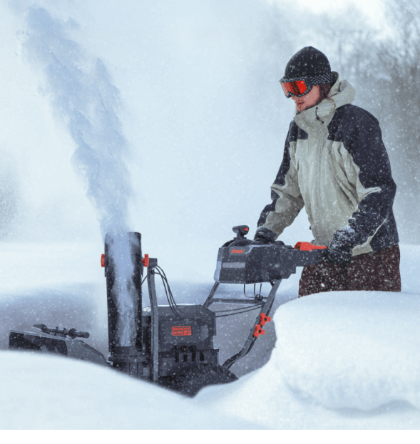 A person wearing winter gear operates a snow blower that is throwing a large plume of snow into the air during a snowfall